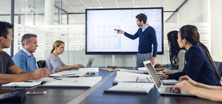 a male professional explains a numbers chart on an overhead screen to coworkers seated at a conference table