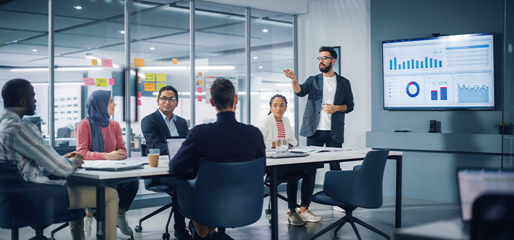 a man talks to coworkers in a conference room with a business chart on the wall