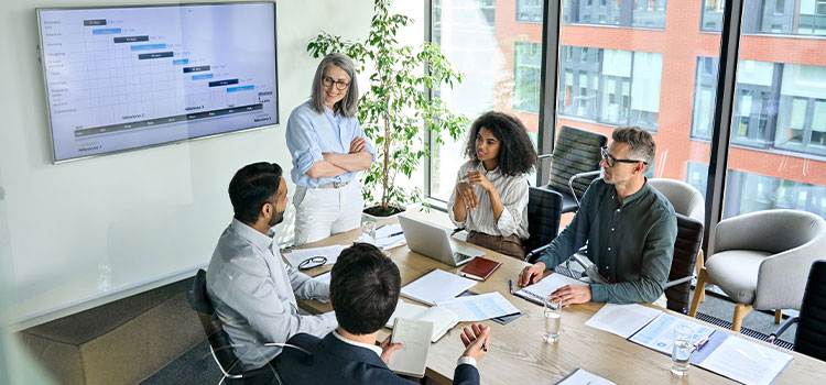 a  woman standing in front of a wall screen with data talks to other professionals gathered around a table