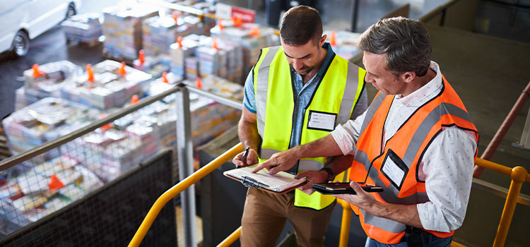 two men discuss information on a clipboard as they survey goods in a warehouse